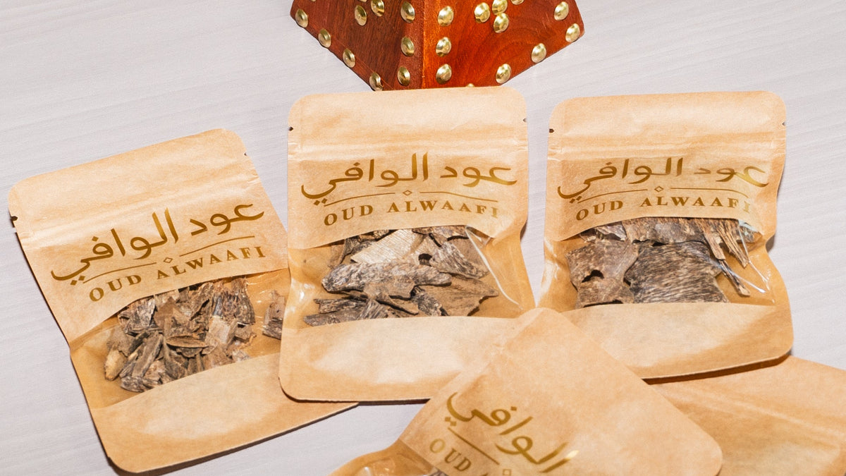 Four small beige resealable pouches labeled QUD ALWAaFI are arranged on a light surface, with dried herbal pieces visible through the front windows. A decorative orange-brown object with gold studs appears at the top center.